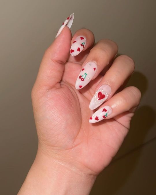 Almond-shaped nails with a soft white base, adorned with tiny red hearts, cherry motifs, and pearl accents valentine's day nails