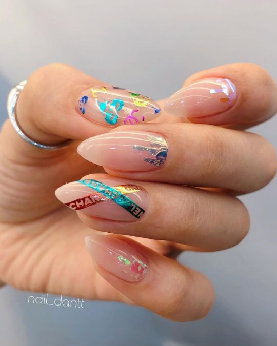 Almond-shaped nails with a nude base, adorned with colorful foil accents and Chanel logo