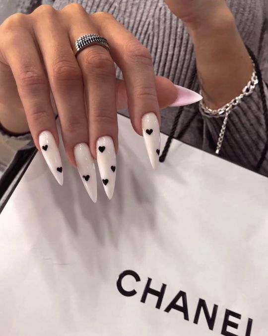 Sharp stiletto nails in pearlescent white, adorned with tiny black hearts