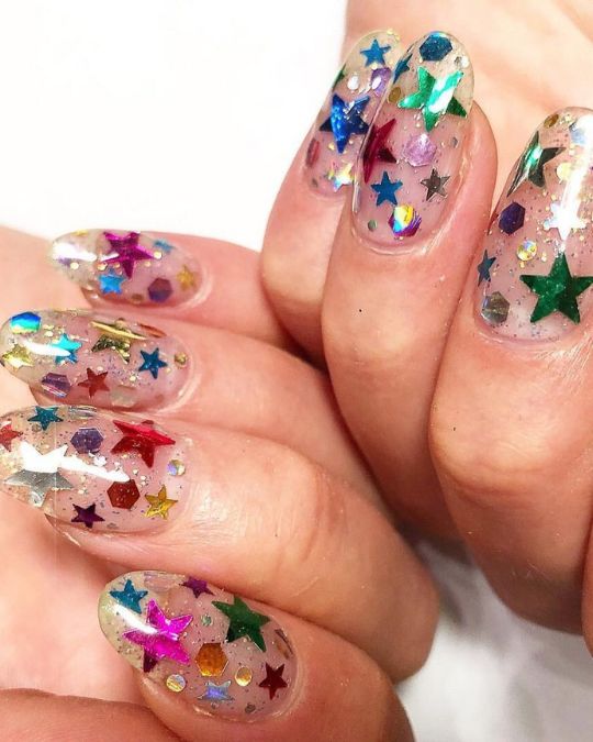 Oval-shaped nails with a translucent pink base, adorned with colorful metallic stars and geometric shapes