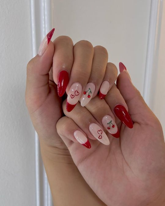Almond-shaped nails showcase a captivating blend of glossy red and soft pink, adorned with delicate hearts and charming cherry motifs