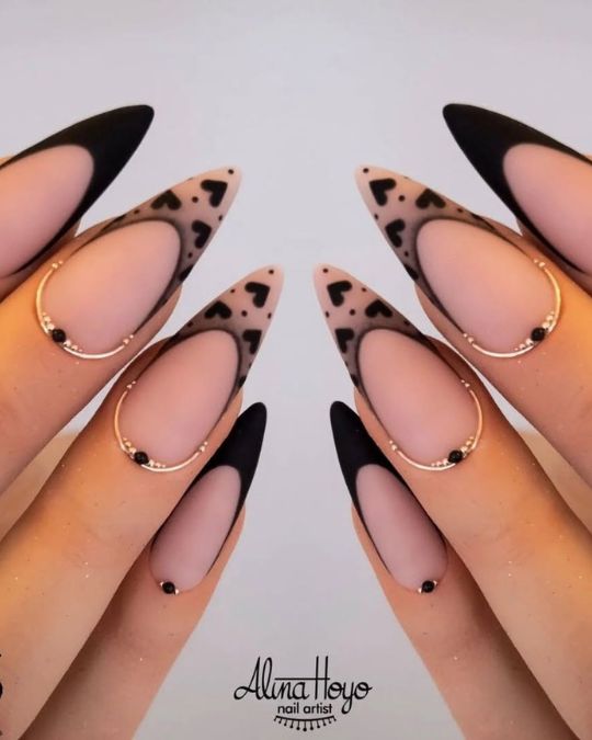 Sharp stiletto nails combine a subtle nude base with striking matte black French tips, accented by delicate heart designs
